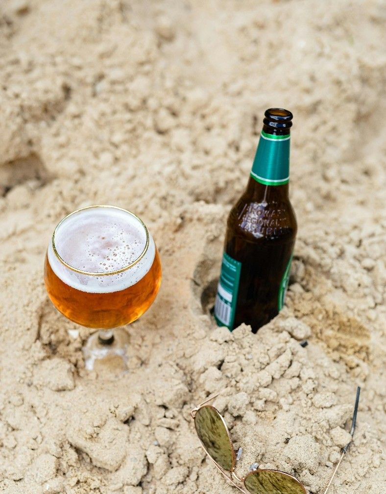 Cold beer in a tulip glass next to a bottle and sunglasses nestled in the sand at Chubby's Bier Garden