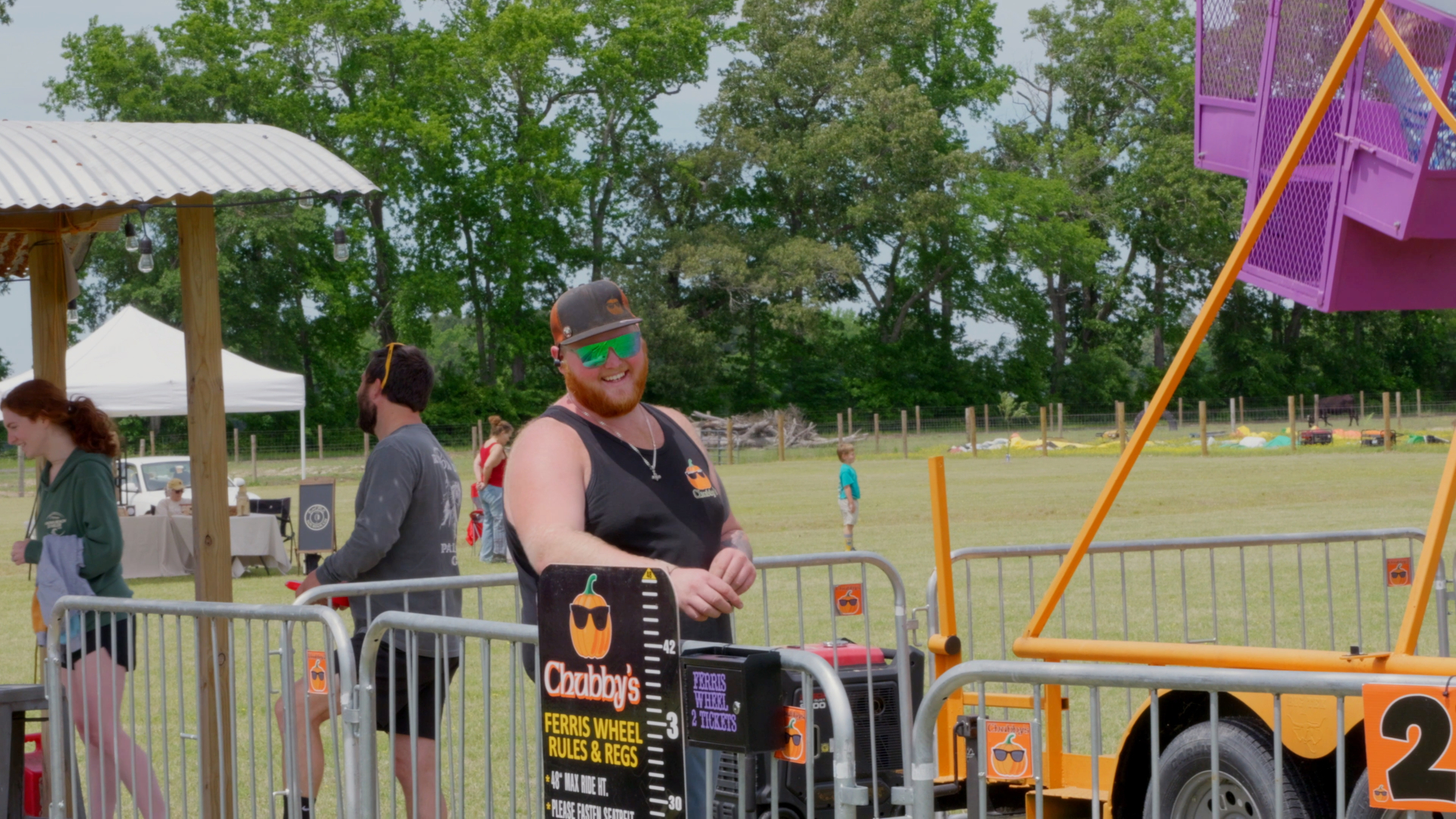 Chubby's Ferris Wheel operator smiling beside the ride's rules-and-regs sign on a sunny day