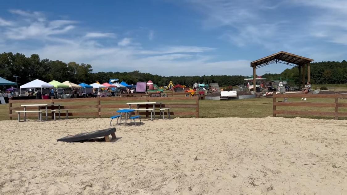 Sandy beach hangout with picnic tables and cornhole, vendor tents and pavilion stage in the background